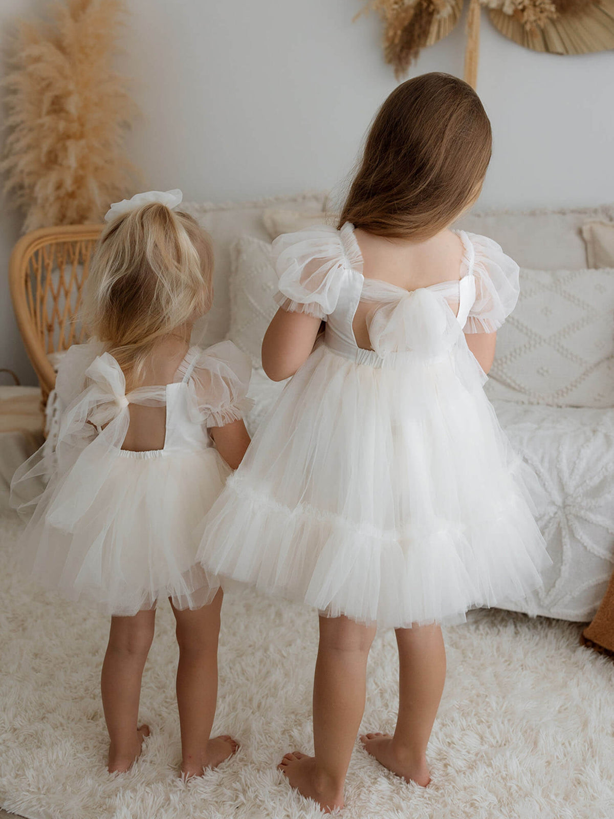 Two sisters stand wearing matching cream Gabrielle flower girl rompers and dresses, showing the tulle bow back.