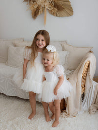 Sisters stand together wearing matching flower girl outfits. One wears our Gabrielle cream flower girl dress and the other wears our Gabrielle cream toddler flower girl romper, along with a matching tulle bow hair clip.