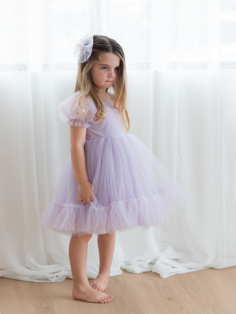 Gabrielle lilac flower girl dress is worn by a young girl. She wears a matching lilac tulle bow in her hair.