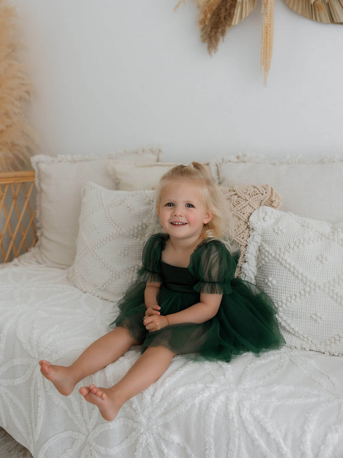 A toddler girl sits on a white couch, wearing a green flower girl dress for babies.