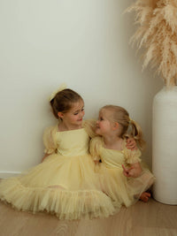 Sisters sit wearing matching yellow flower girl outfits. The older sister wears our Gabrielle lemon flower girl dress, and the younger our Gabrielle lemon baby flower girl romper.
