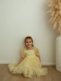 A young girl sits on the floor wearing our Gabrielle lemon yellow party dress and matching lemon tulle hair bow.