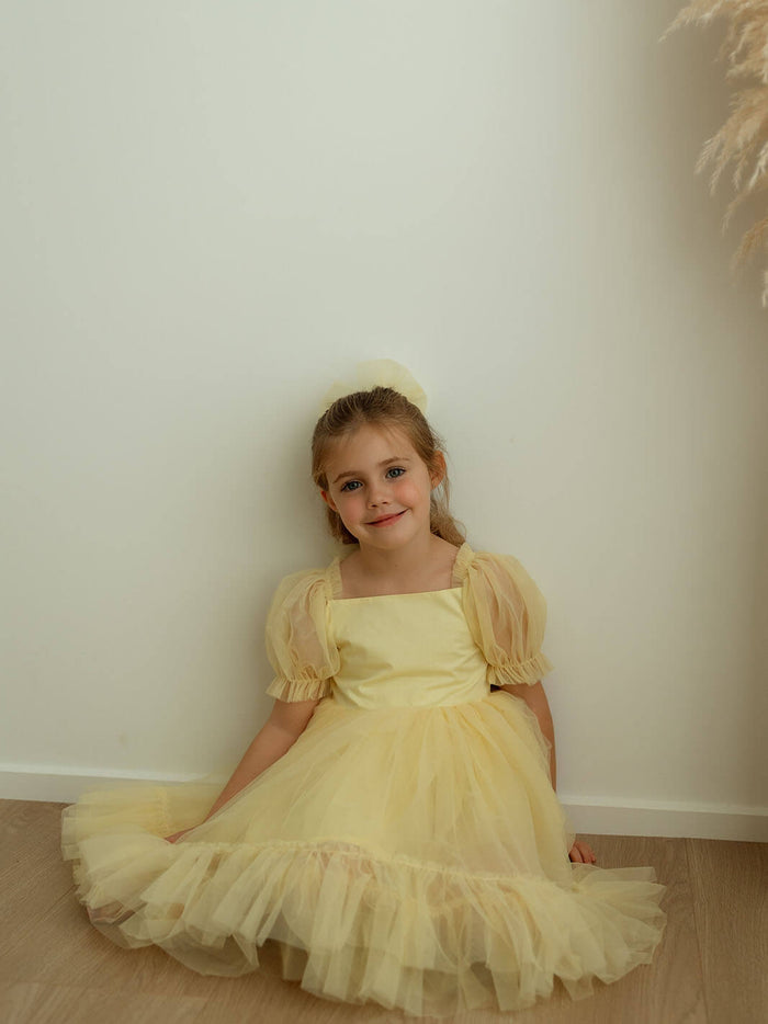 A little girl sits wearing our Gabrielle lemon yellow flower girl dress with tulle puff sleeves.