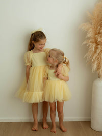 Sisters stand wearing matching lemon yellow flower girl outfits. The older sister wears our Gabrielle lemon flower girl dress and the younger sister wears our Gabrielle lemon flower girl romper.
