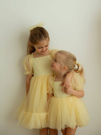 Young sisters stand hugging wearing matching lemon Gabrielle flower girl outfits.