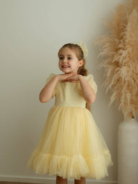 A little girl stands wearing our Gabrielle lemon yellow flower girl dress and matching yellow tulle bow.