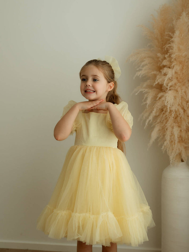 A little girl stands wearing our Gabrielle lemon yellow flower girl dress and matching yellow tulle bow.
