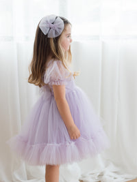 The Gabrielle pastel flower girl dress in lilac, shown from the side on a young girl, along with a matching lilac tulle hair bow.