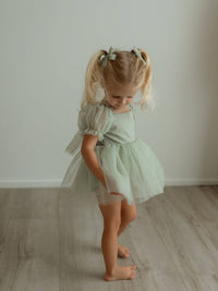 Sage green baby flower girl romper, our Gabrielle romper, is worn by a toddler. She also wears matching sage green tulle pigtail bows.