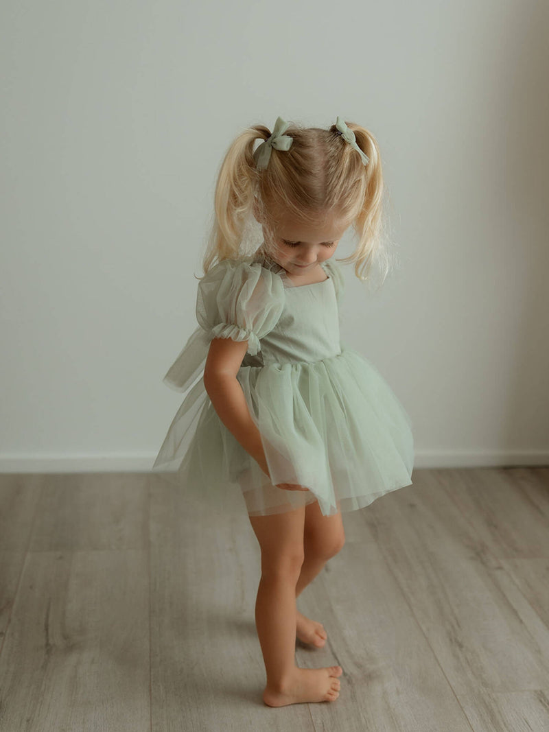 Sage green baby flower girl romper, our Gabrielle romper, is worn by a toddler. She also wears matching sage green tulle pigtail bows.
