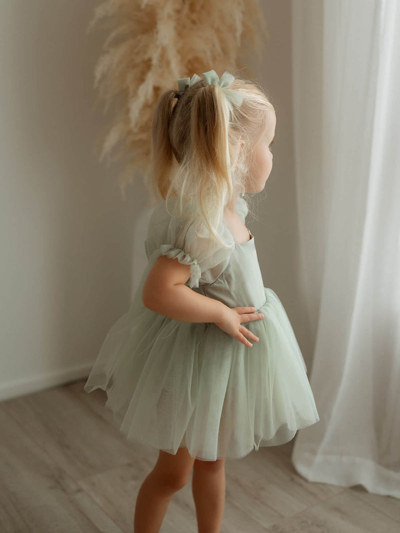 A little girl looks out of a window wearing our Gabrielle sage green baby flower girl romper and matching tulle pigtail bows.