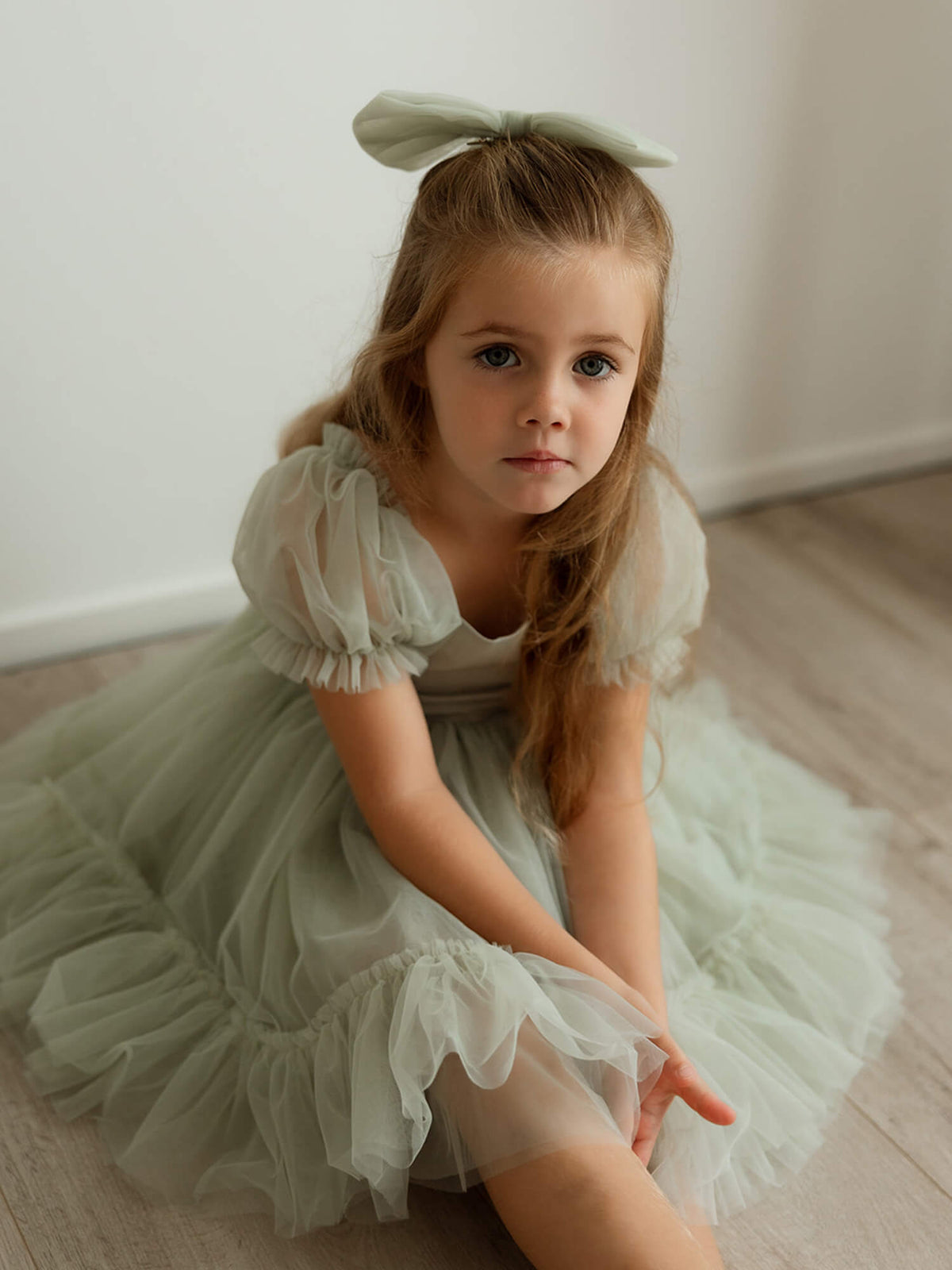Sage green tulle flower girl dress, our Gabrielle dress, is worn by a young girl who sits on the floor.