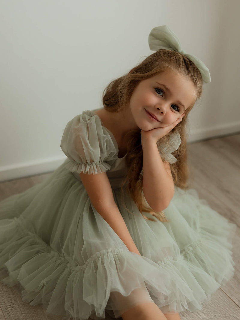 A little girl smiles with her head resting on her hand. She is wearing our Gabrielle sage tulle flower girl dress and matching large tulle bow in sage.