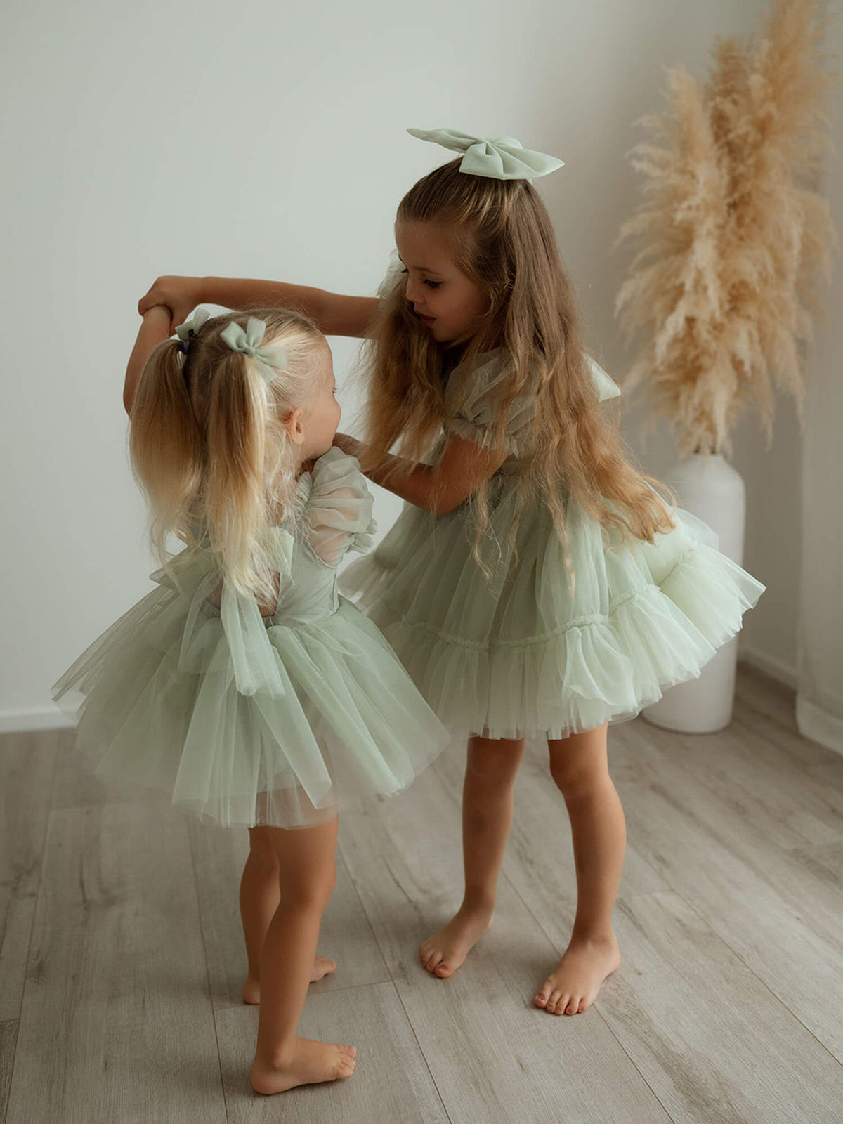 Sisters hold hands dancing wearing our Gabrielle flower girl romper and dress in sage green tulle.