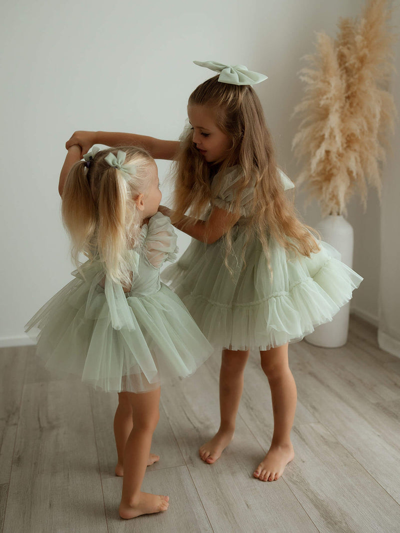 Sisters hold hands dancing wearing our Gabrielle flower girl romper and dress in sage green tulle.