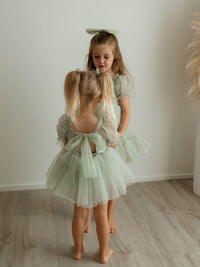The back of our Gabrielle baby flower girl romper in sage green, worn by a toddler. Her sister wears our matching Gabrielle flower girl dress in sage green.