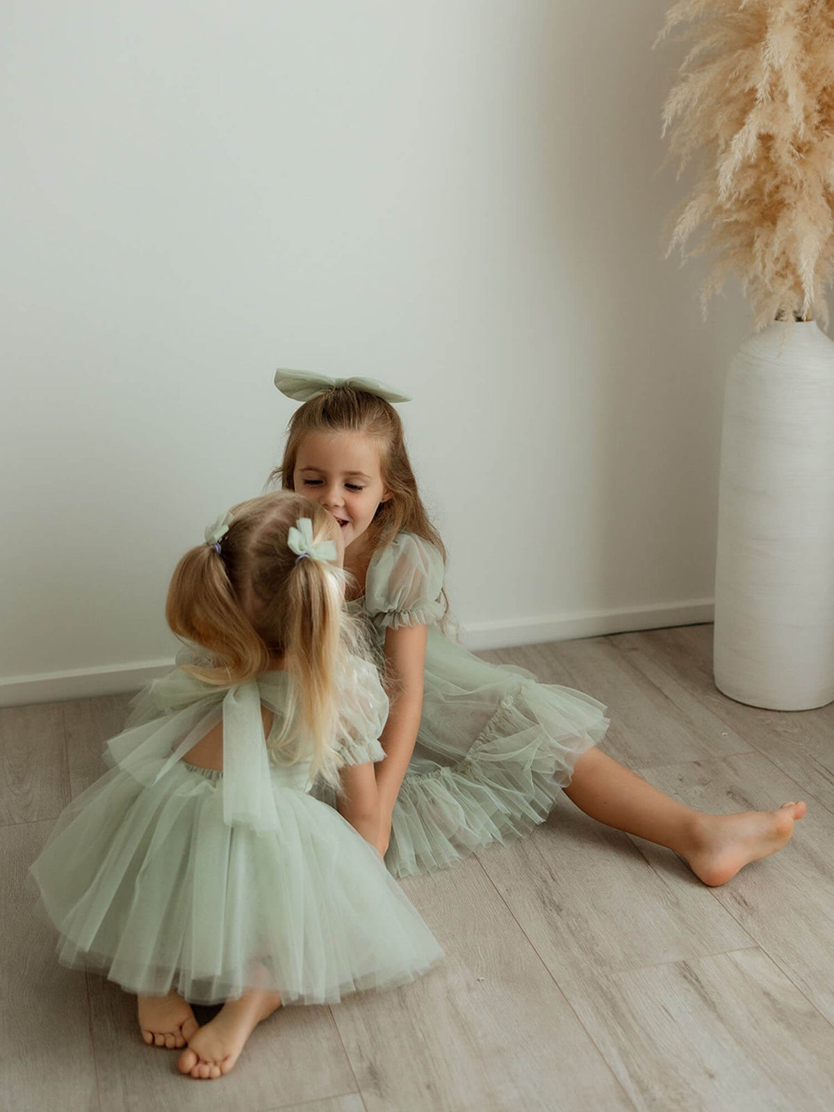 Sisters sit on the floor together, wearing our Gabrielle sage baby flower girl romper and Gabrielle sage flower girl dress.