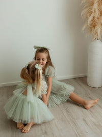 Sisters sit on the floor together, wearing our Gabrielle sage baby flower girl romper and Gabrielle sage flower girl dress.