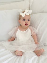 A baby sits wearing our Gia baby flower girl romper and an ivory bow headband.