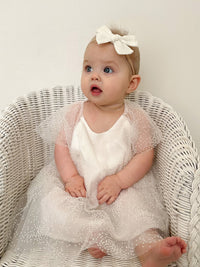 A baby girl sits in a small chair, wearing our Gia baby flower girl romper and an ivory bow in her hair.