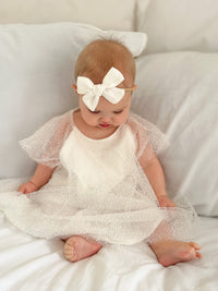 A baby sits on a bed wearing our Gia baby flower girl dress, made from flocked confetti tulle.