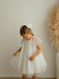 A little girl stands wearing our Gia flower girl dress and matching ivory confetti tulle bow.
