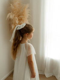 A little girl stands near a window wearing our Gia flower girl dress in flocked dotted tulle and a matching tulle bow hairpiece. 