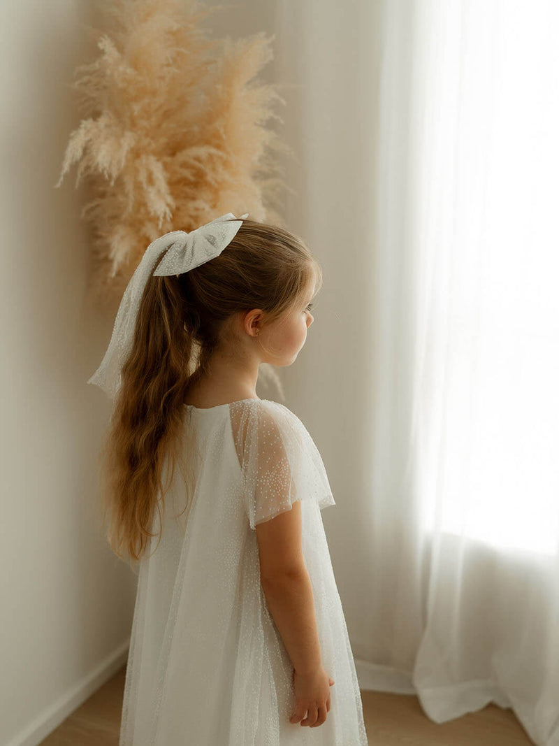 A little girl stands near a window wearing our Gia flower girl dress in flocked dotted tulle and a matching tulle bow hairpiece. 