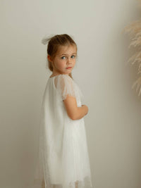 Young girl in a white flower girl dress dress with flocked details against a plain background.