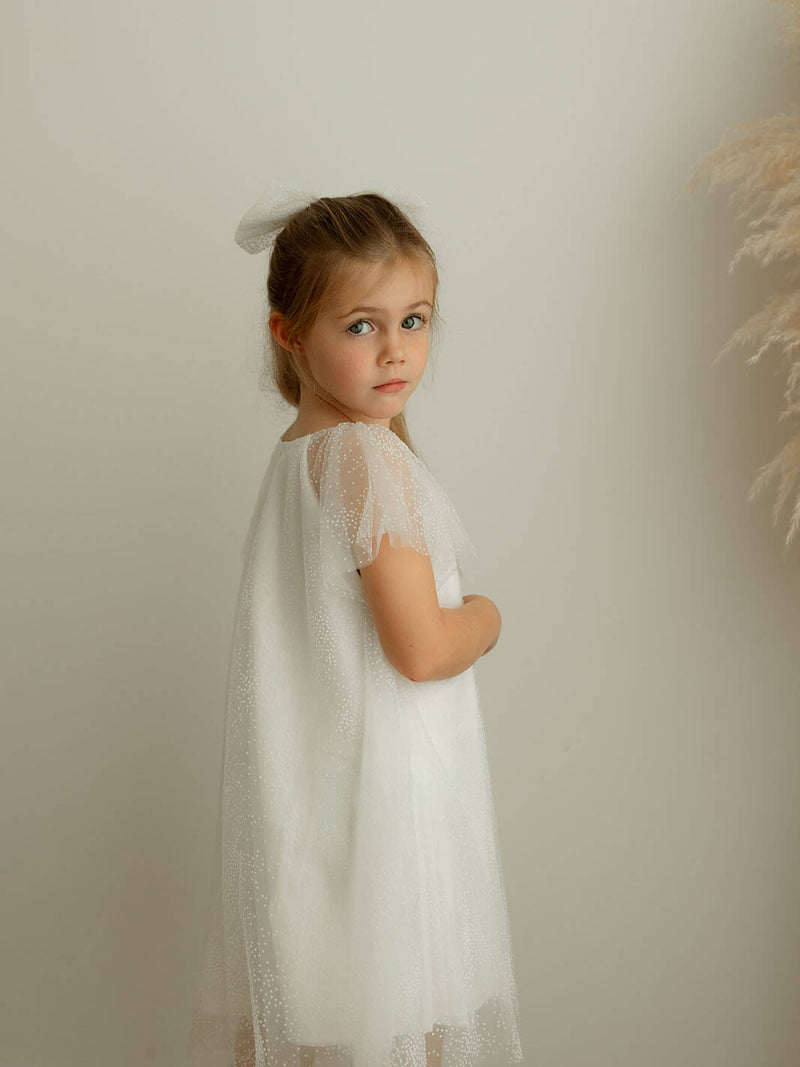 Young girl in a white flower girl dress dress with flocked details against a plain background.