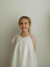 Young girl smiles wearing an ivory flower girl dress with sheer sleeves.