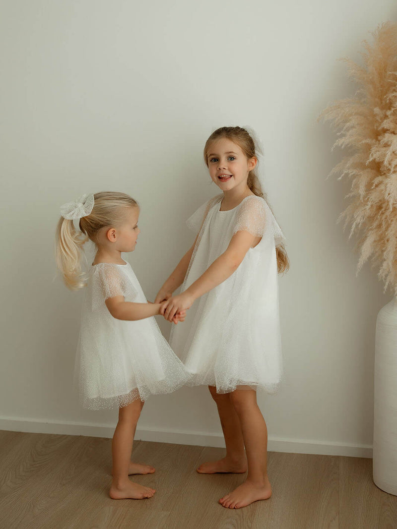 Two young girls in white flower girl dresses holding hands, wearing matching tulle bows in their hair.