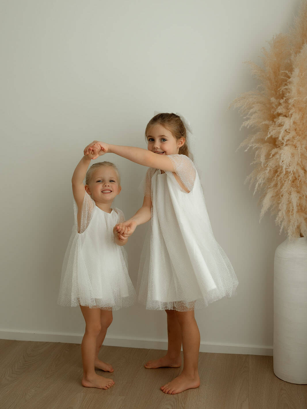 Two young girls in white flower girl dresses holding hands against a plain wall with a vase of dried plants.
