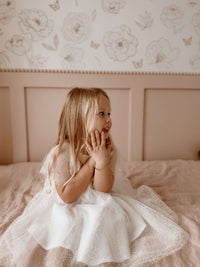 A young girl sits on a bed, wearing our Gia flocked dot tulle flower girl dress.