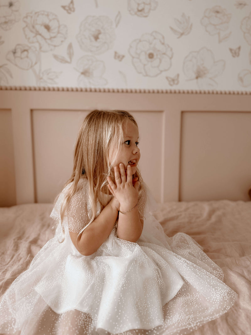 A young girl sits on a bed, wearing our Gia flocked dot tulle flower girl dress.