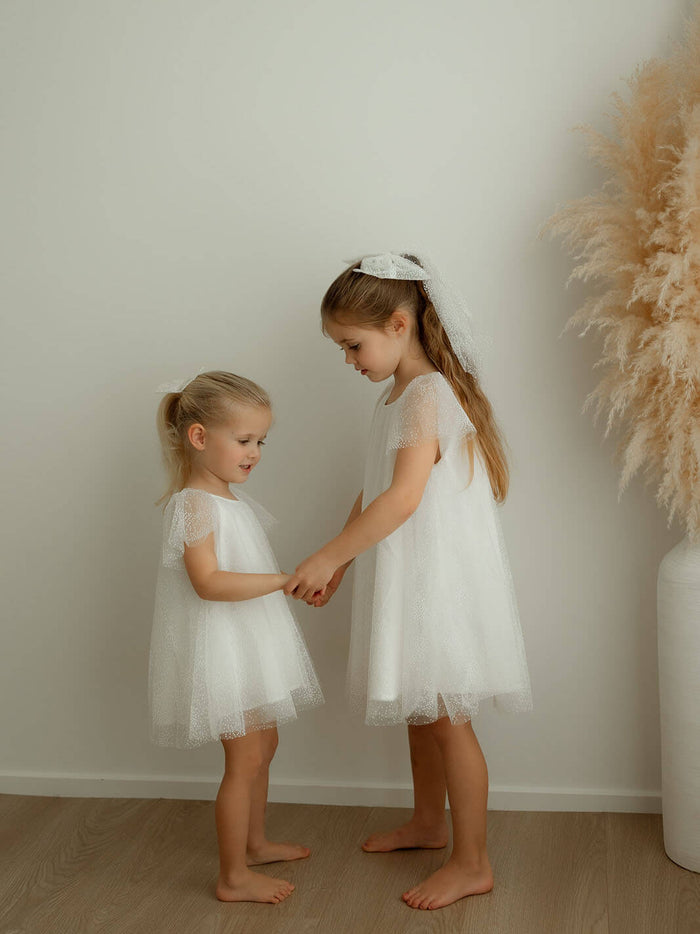 Sisters stand holding hands, one wears our Gia ivory baby flower girl romper and the other wears our Gia flower girl dress.