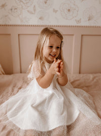 Young girl in our Gia flower girl dress, sitting on a bed with a floral headboard.