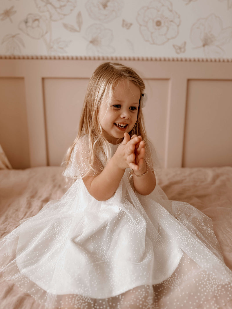 Young girl in our Gia flower girl dress, sitting on a bed with a floral headboard.