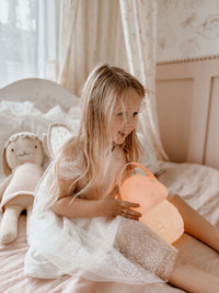 Young girl in our Gia flower girl dress, holding a pink lamp on a bed with a plush toy.