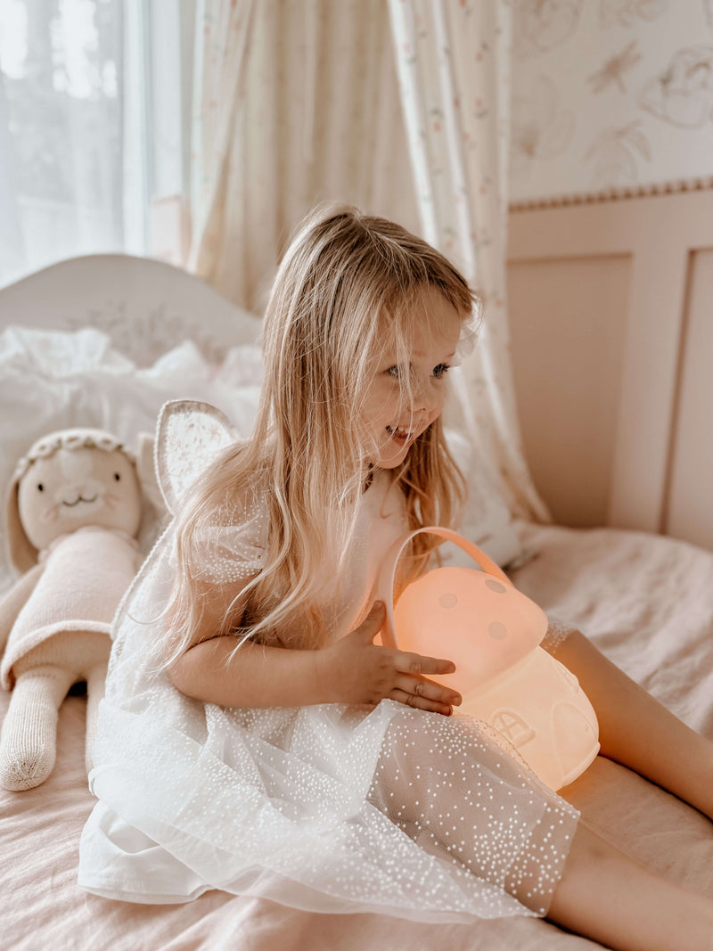 Young girl in our Gia flower girl dress, holding a pink lamp on a bed with a plush toy.