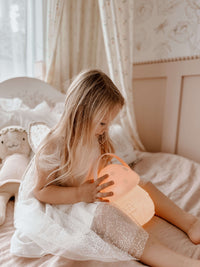 Child wears our Gia flower girl dress with confetti tulle, holding a glowing lamp in a cozy bedroom.