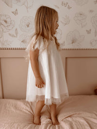 Young girl in our Gia flower girl dress, standing on a bed with floral wallpaper in the background.