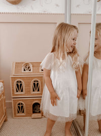 Young girl in our Gia flower girl dress, standing next to a wooden dollhouse in a room.