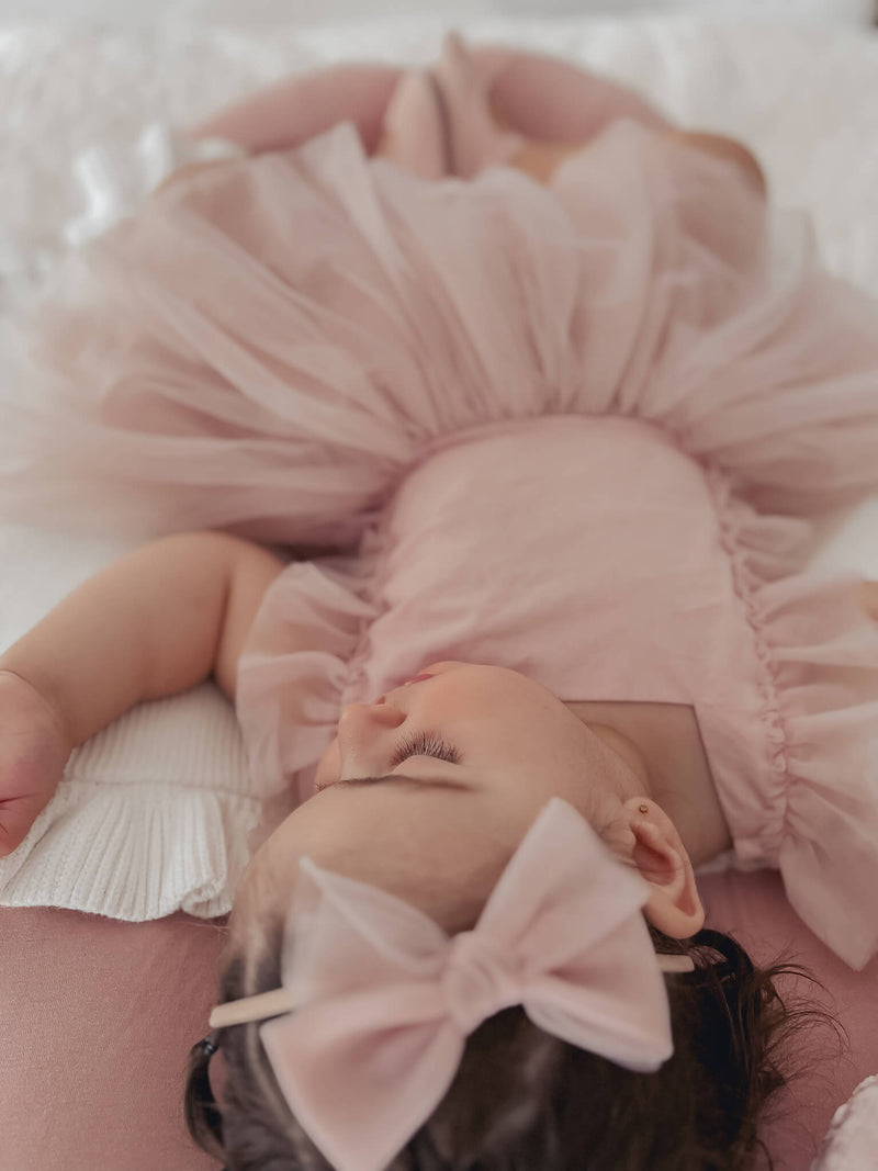 Gigi dusty pink baby dress is worn by a young baby girl who is asleep. She also wears our dusty pink tulle bow headband.lep