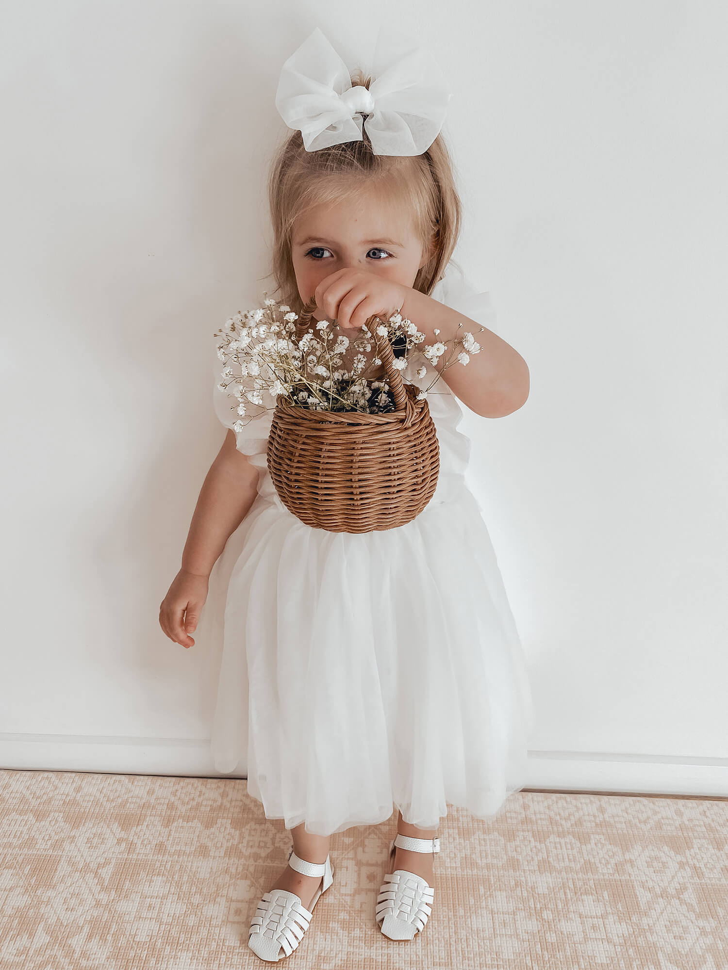 A young flower girl holds a basket of flowers while wearing our Gigi flower girl dress in ivory, and an ivory tulle bow in her hair.