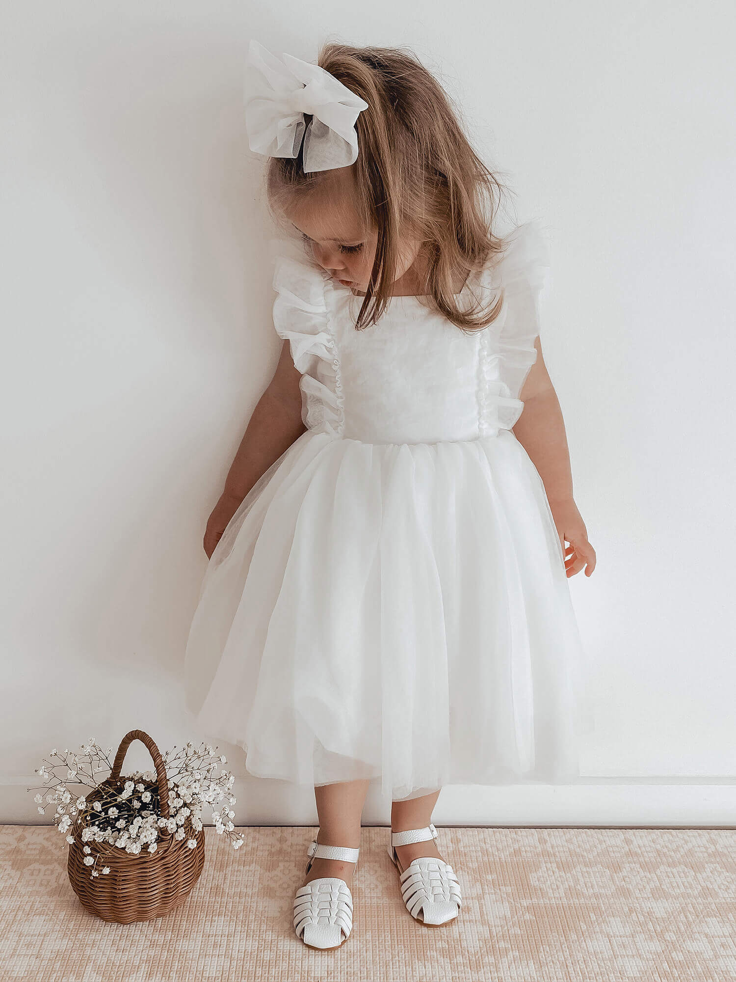 Gigi white flower girl dress worn by a young girl, she also wears our ivory tulle bow in her hair and stands beside a basket of flowers.