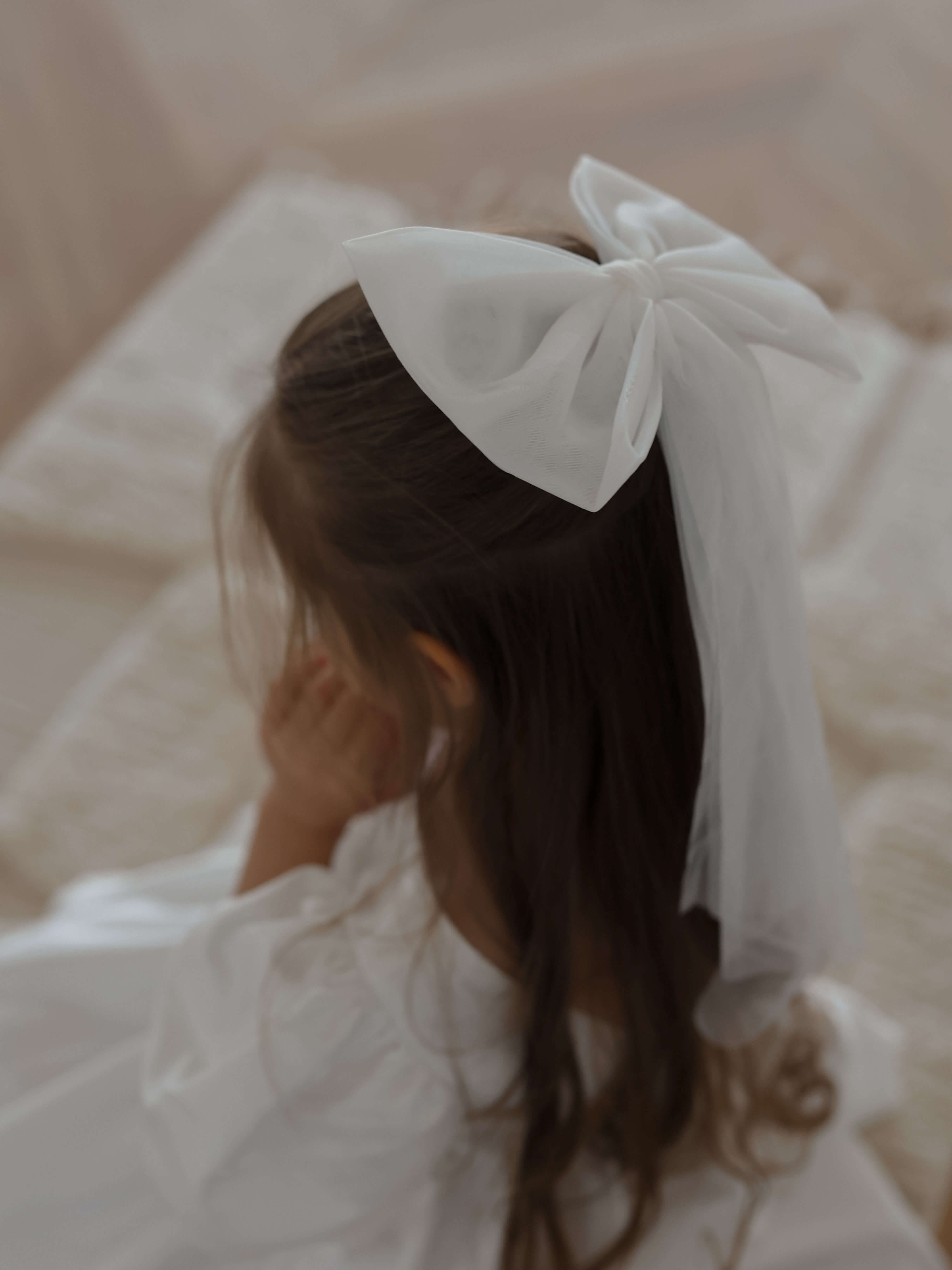 Large tulle bow hair clip in ivory is worn by a young girl. She also wears a satin flower girl dress.