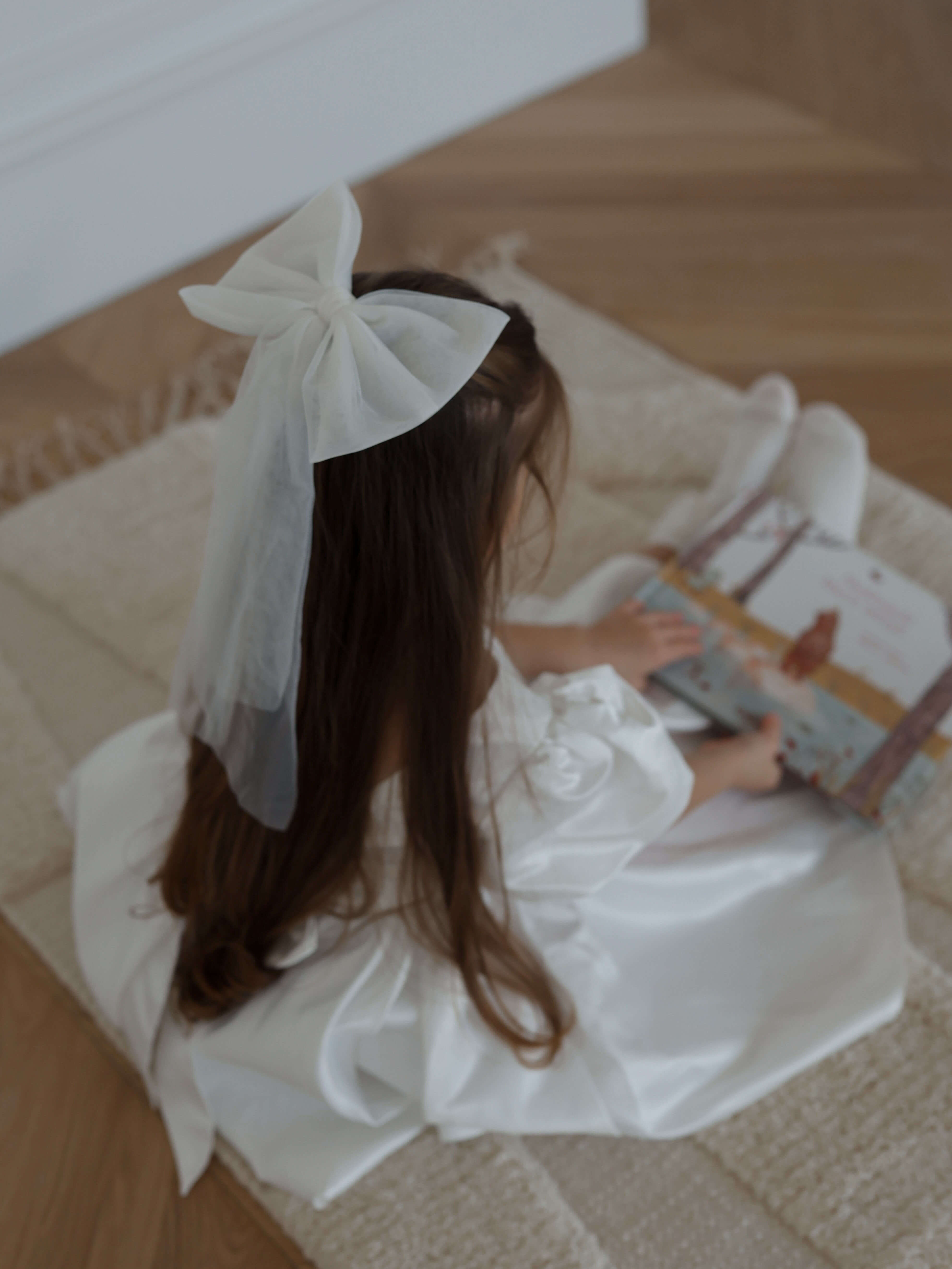 A young girl sits reading, she wears a satin flower girl dress and our large tulle bow in ivory in her hair.
