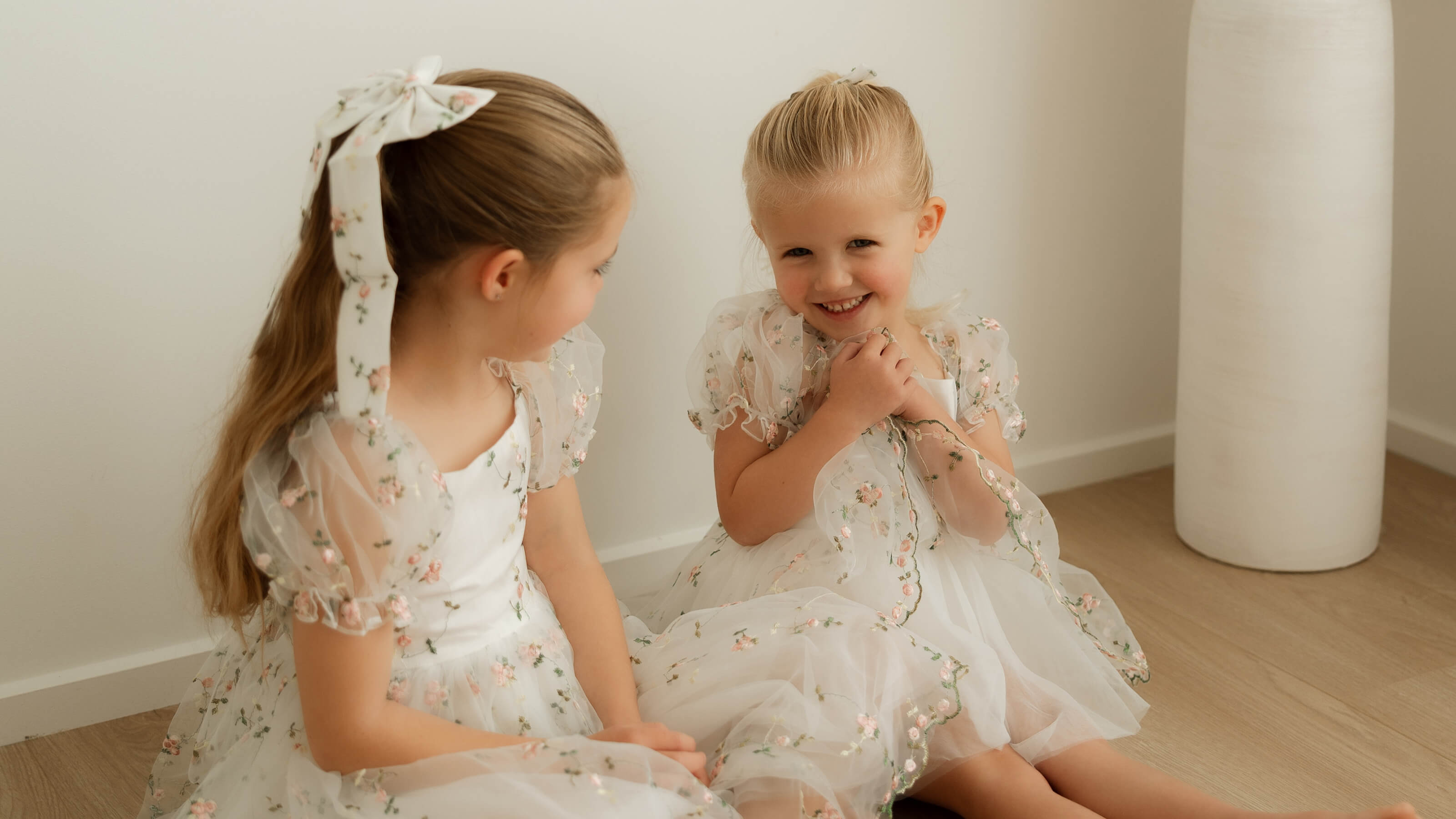 Young girls sit wearing matching floral special occasion dresses.