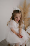A young girl smiles wearing our Gracie puff sleeve flower girl dress in ivory and tulle bow hair clip.